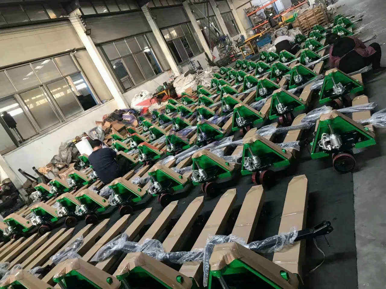 Xe nâng tay thủy lực màu xanh xếp hàng trong kho, công nhân đang lắp ráp và đóng gói sản phẩm